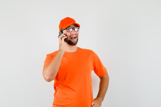 Repartidor en camiseta naranja, gorra hablando por teléfono móvil y mirando feliz, vista frontal.