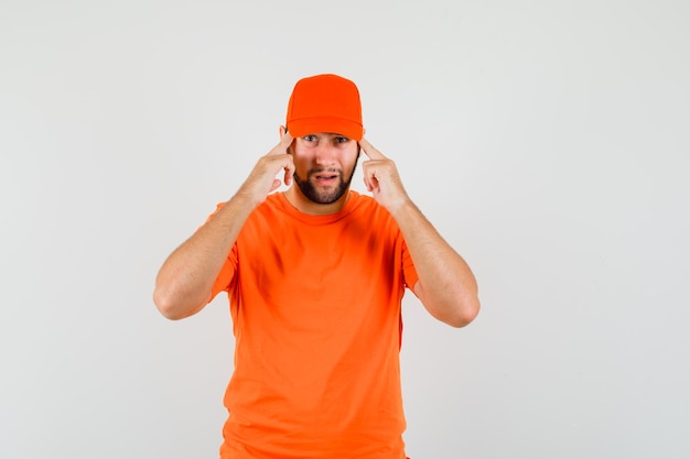 Repartidor en camiseta naranja, gorra manteniendo los dedos en las sienes y luciendo sensato, vista frontal.