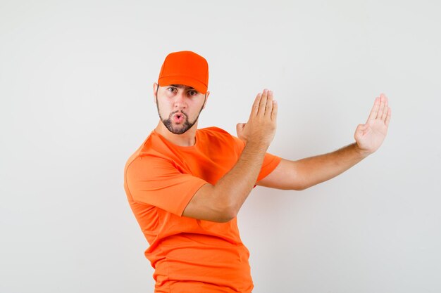 Repartidor en camiseta naranja, gorra mostrando gesto de tajo de karate y mirando confiado, vista frontal.