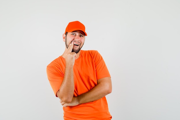 Repartidor en camiseta naranja, gorra que muestra un diente dolorido y que parece incómodo, vista frontal.