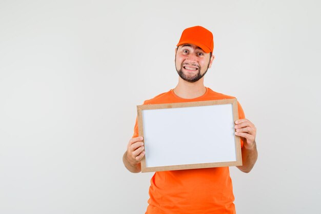 Repartidor en camiseta naranja, gorra sosteniendo un marco en blanco y mirando alegre, vista frontal.