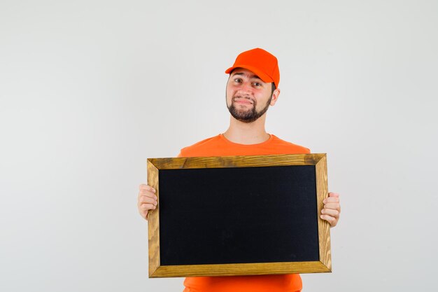Repartidor en camiseta naranja, gorra sosteniendo una pizarra en blanco y mirando suave, vista frontal.