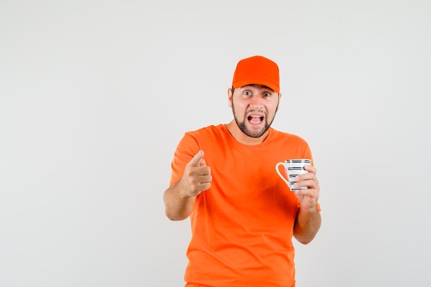 Repartidor en camiseta naranja, gorra sosteniendo una taza de bebida, apuntando y mirando desconcertado, vista frontal.