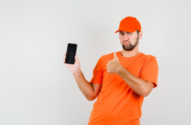 Repartidor en camiseta naranja, gorra sosteniendo el teléfono móvil con el pulgar hacia arriba y mirando triste, vista frontal.