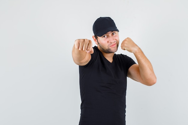 Foto gratuita repartidor en camiseta negra, gorra de pie en pose de boxeador y mirando confiado