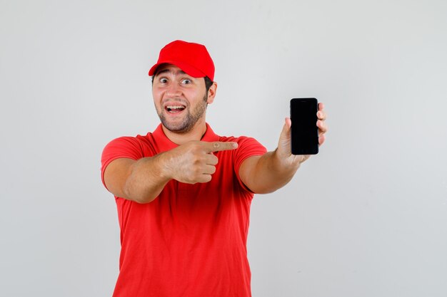 Repartidor en camiseta roja, gorra apuntando al teléfono inteligente y mirando alegre