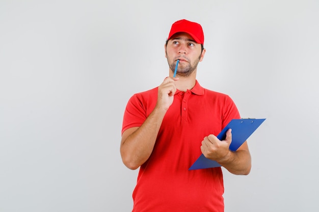 Repartidor en camiseta roja, gorra mirando hacia arriba con lápiz y portapapeles y mirando pensativo