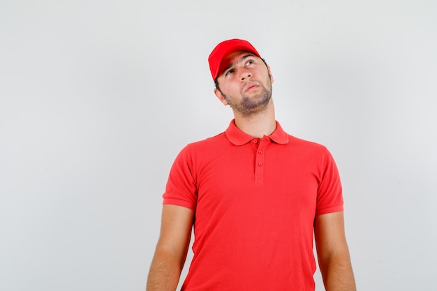 Repartidor en camiseta roja, gorra mirando hacia arriba y mirando pensativo