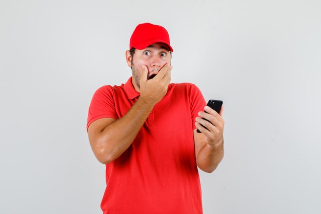 Repartidor en camiseta roja, gorra sosteniendo el teléfono inteligente con la mano en la boca y mirando sorprendido