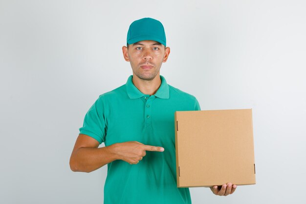 Repartidor en camiseta verde y gorra mostrando caja de cartón con el dedo