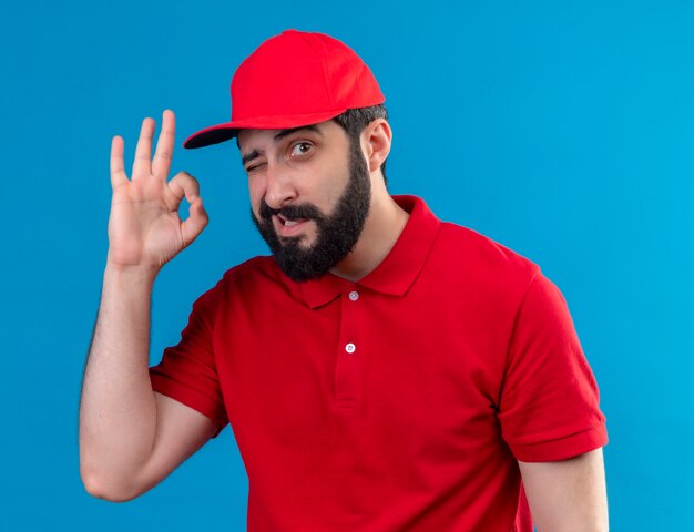 Repartidor caucásico guapo joven confiado con uniforme rojo y gorra guiñando un ojo y haciendo bien signo aislado en azul