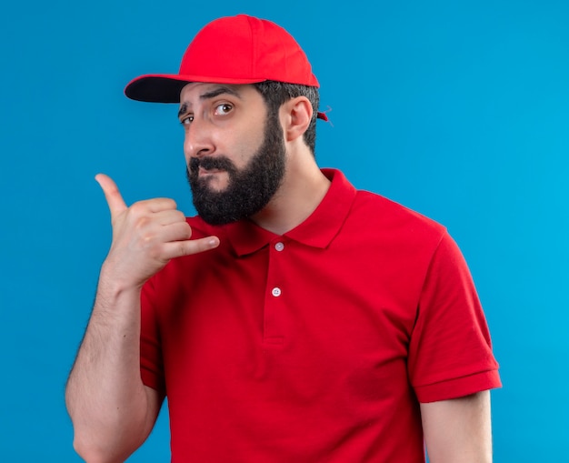 Repartidor caucásico guapo joven confiado con uniforme rojo y gorra haciendo gesto de llamada aislado en azul