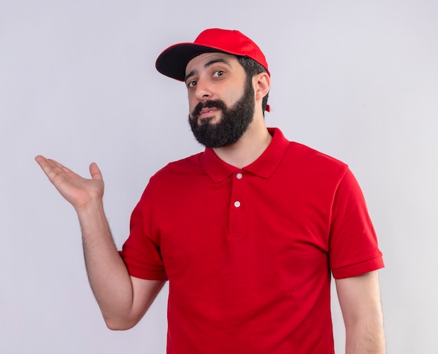 Repartidor caucásico guapo joven confiado con uniforme rojo y gorra mostrando la mano vacía aislada en blanco