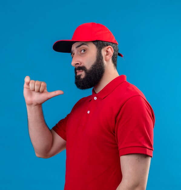 Repartidor caucásico guapo joven confiado con uniforme rojo y gorra de pie en la vista de perfil apuntando a sí mismo aislado en azul