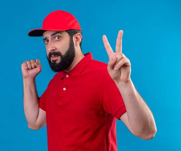 Repartidor caucásico guapo joven confiado con uniforme rojo y gorra de pie en la vista de perfil y haciendo el signo de la paz y apretando el puño aislado en azul