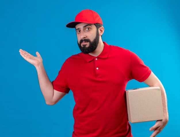 Repartidor caucásico guapo joven confiado con uniforme rojo y gorra sosteniendo la caja de cartón y mostrando la mano vacía aislada en azul