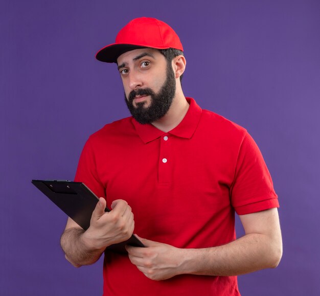 Repartidor caucásico guapo joven confiado con uniforme rojo y gorra sosteniendo portapapeles aislado en púrpura