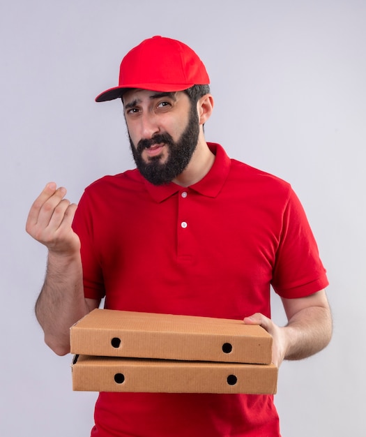 Repartidor caucásico guapo joven vestido con uniforme rojo y gorra sosteniendo cajas de pizza y gesticulando dinero mirando a cámara aislada sobre fondo blanco