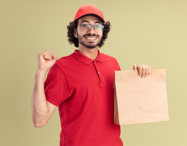 Repartidor caucásico joven alegre en uniforme rojo y gorra con gafas sosteniendo el paquete de papel haciendo un gesto fuerte