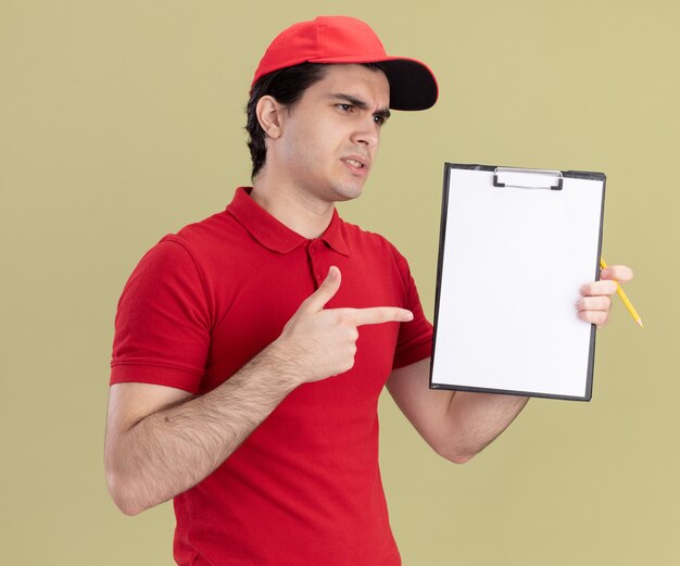 Repartidor caucásico joven con el ceño fruncido en uniforme rojo y gorra que muestra el portapapeles