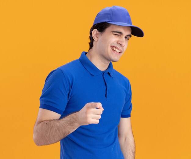 Repartidor caucásico joven confiado en uniforme azul y gorra guiñando un ojo mirando y apuntando a la cámara solated sobre fondo naranja