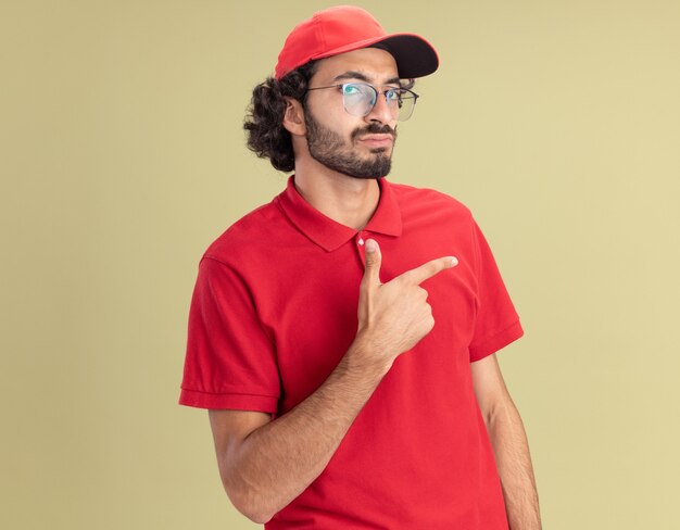 Repartidor caucásico joven confiado en uniforme rojo y gorra con gafas apuntando al lado