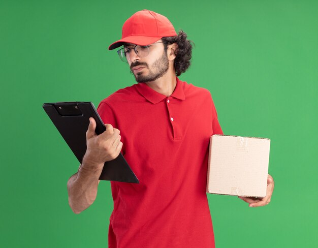 Repartidor caucásico joven confuso en uniforme rojo y gorra con gafas sosteniendo cardbox y portapapeles mirando el portapapeles