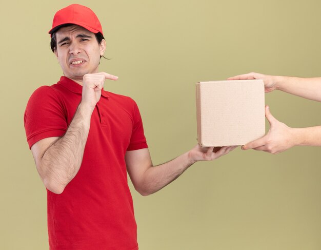 Repartidor caucásico joven disgustado con uniforme rojo y gorra dando caja de cartón al cliente apuntando a la caja
