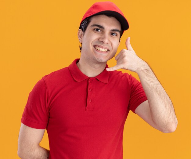 Repartidor caucásico joven sonriente en uniforme azul y gorra haciendo gesto de llamada aislado en la pared naranja