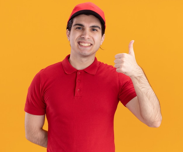 Repartidor caucásico joven sonriente en uniforme azul y gorra manteniendo la mano detrás de la espalda mostrando el pulgar hacia arriba