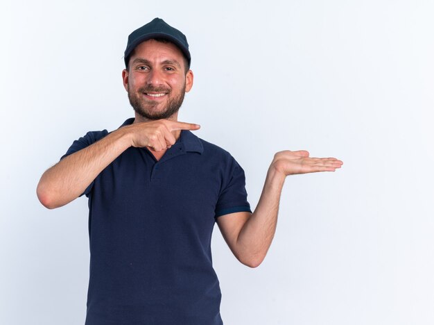 Repartidor caucásico joven sonriente en uniforme azul y gorra mirando a la cámara mostrando la mano vacía apuntando al lado aislado en la pared blanca