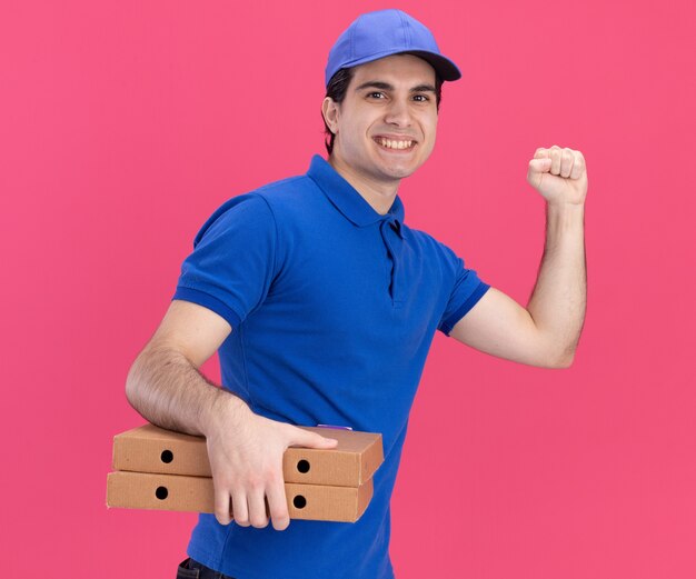 Repartidor caucásico joven sonriente en uniforme azul y gorra de pie en la vista de perfil sosteniendo paquetes de pizza haciendo gesto de golpe aislado en la pared rosa