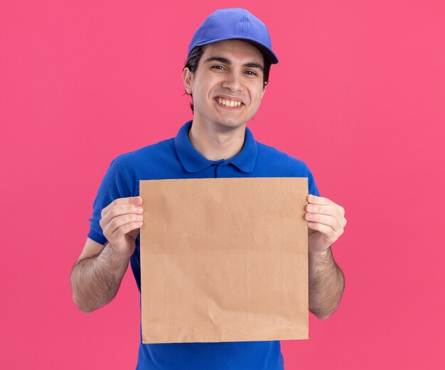 Repartidor caucásico joven sonriente en uniforme azul y gorra sosteniendo el paquete de papel