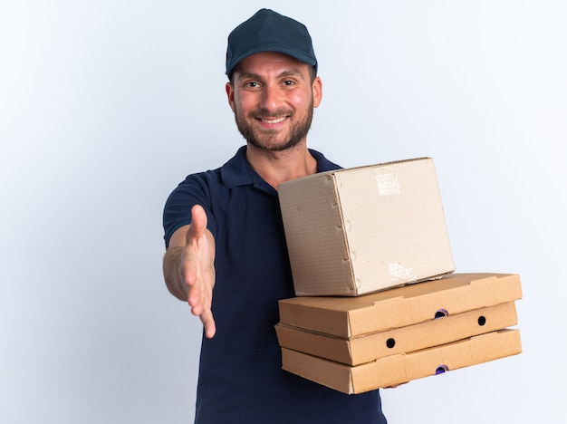 Repartidor caucásico joven sonriente en uniforme azul y gorra sosteniendo paquetes de pizza y caja de cartón mirando a cámara haciendo gesto de saludo aislado en la pared blanca con espacio de copia