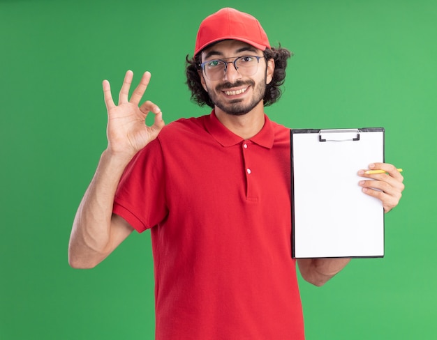 Repartidor caucásico joven sonriente en uniforme rojo y gorra con gafas mostrando el portapapeles a la cámara haciendo bien firmar con lápiz en mano