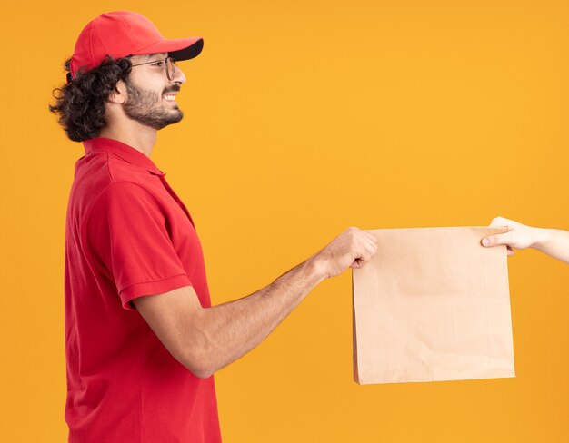 Repartidor caucásico joven sonriente en uniforme rojo y gorra con gafas de pie en la vista de perfil dando paquete de papel al cliente mirando al cliente