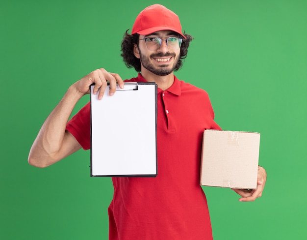 Repartidor caucásico joven sonriente en uniforme rojo y gorra con gafas sosteniendo una caja de cartón y mostrando el portapapeles