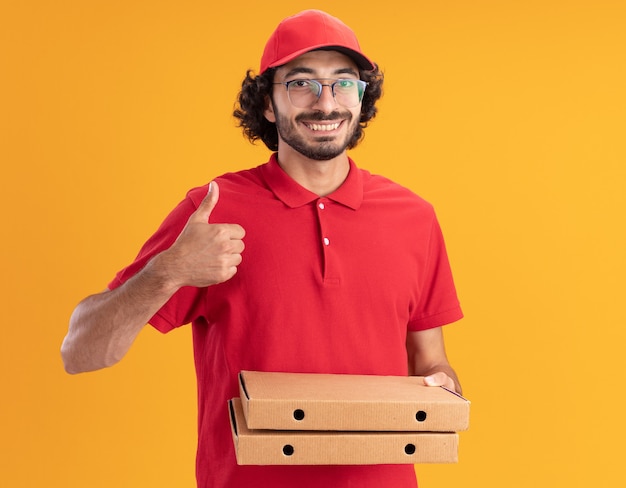 Repartidor caucásico joven sonriente en uniforme rojo y gorra con gafas sosteniendo paquetes de pizza mostrando el pulgar hacia arriba aislado en la pared naranja con espacio de copia