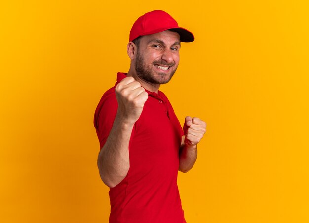 Repartidor caucásico joven sonriente en uniforme rojo y gorra de pie en la vista de perfil mirando a la cámara haciendo gesto de boxeo aislado en la pared naranja con espacio de copia