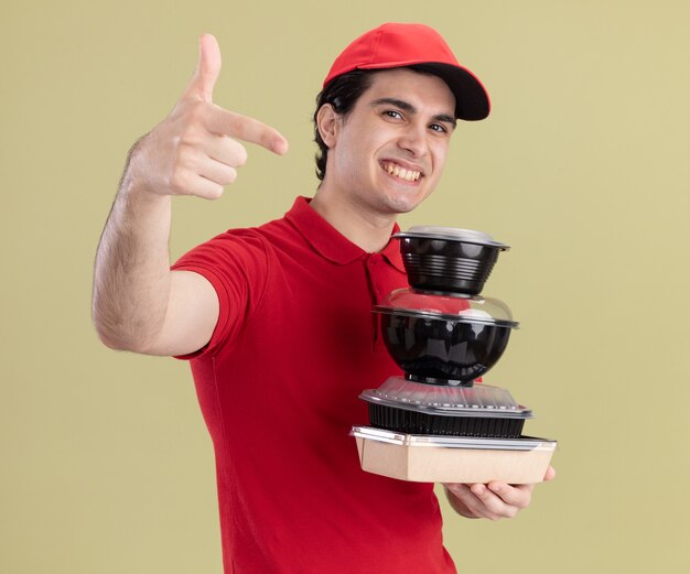 Repartidor caucásico joven sonriente en uniforme rojo y gorra sosteniendo contenedores de comida y paquete de comida de papel mirando y apuntando aislado en la pared verde oliva