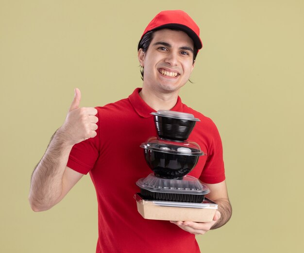 Repartidor caucásico joven sonriente en uniforme rojo y gorra sosteniendo contenedores de comida y paquete de comida de papel mostrando el pulgar hacia arriba aislado en la pared verde oliva