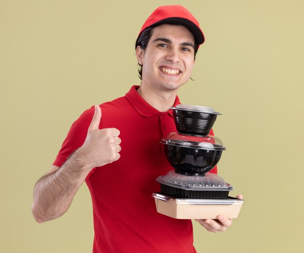 Repartidor caucásico joven sonriente en uniforme rojo y gorra sosteniendo envases de comida y paquete de comida de papel mostrando el pulgar hacia arriba