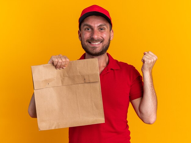 Repartidor caucásico joven sonriente en uniforme rojo y gorra sosteniendo el paquete de papel haciendo gesto de sí