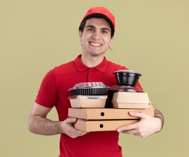 Repartidor caucásico joven sonriente en uniforme rojo y gorra sosteniendo paquetes de pizza con envases de comida y paquetes de comida de papel en ellos aislados en la pared verde oliva