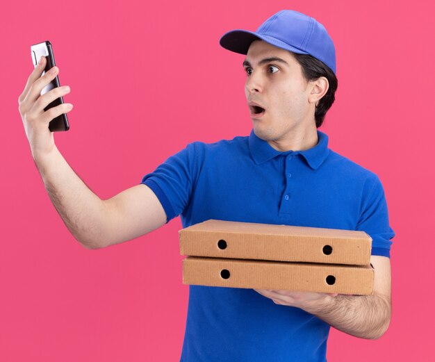 Repartidor caucásico joven sorprendido en uniforme azul y gorra sosteniendo paquetes de pizza estirándose y mirando el teléfono móvil