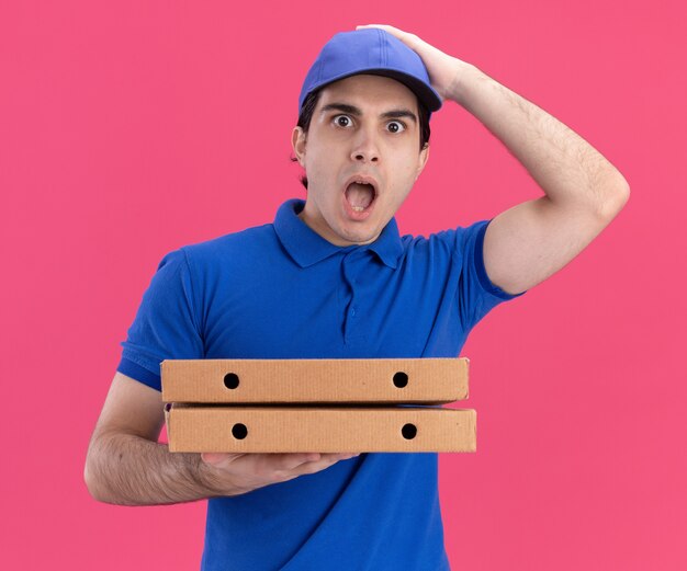 Repartidor caucásico joven sorprendido en uniforme azul y gorra sosteniendo paquetes de pizza poniendo la mano en la cabeza aislada en la pared rosa