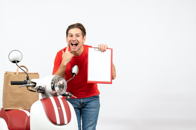 Repartidor feliz joven en uniforme rojo de pie cerca de scooter sosteniendo el documento en la pared blanca