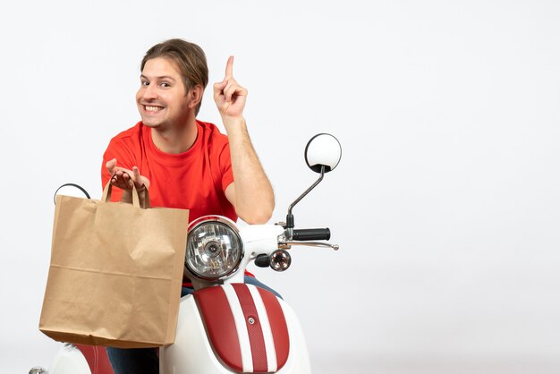 Repartidor feliz joven en uniforme rojo sentado en scooter sosteniendo una bolsa de papel apuntando hacia arriba en la pared blanca