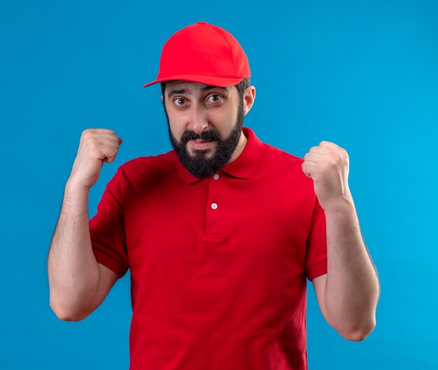 Repartidor guapo joven complacido con uniforme rojo y gorra levantando puños aislados en la pared azul