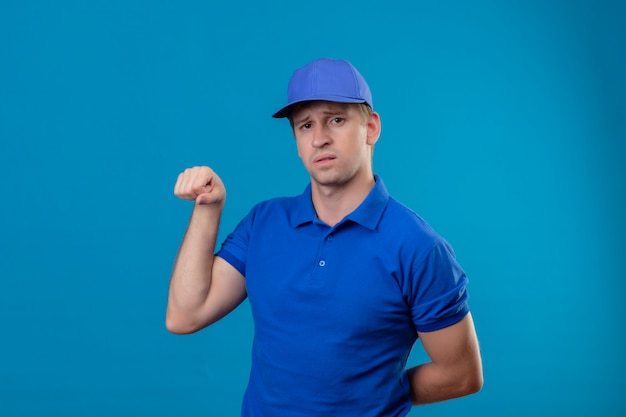 Repartidor guapo joven en uniforme azul y gorra apretando el puño mostrando bíceps mirando confiado parado sobre la pared azul
